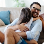 Father laughs as his daughter hugs him while they sit on the living room floor. 